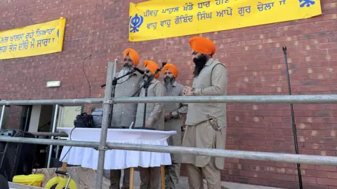 Jules Singh Four men dressed in beige suits and orange turbans speak from microphones outside the gurdwara