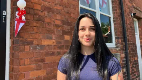 Mairead Smyth/BBC A young woman will long, straight black hair and wearing a purple t-shirt smiles at the camera outside a terraced house decorated with England flags