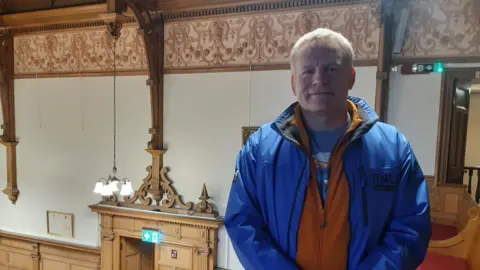 A man with white hair, standing in a council chamber. He is wearing a blue jacket and an orange jacket underneath.