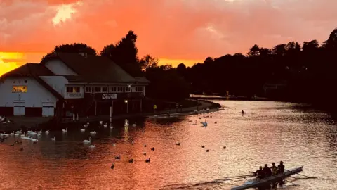 A bright red and orange sky reflect on the Thames in Caversham, with multiple swans and a canoe seen on the water