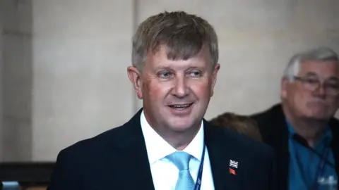 Peter Harris, the leader of Essex County Council, speaking in the council chamber. He is wearing a suit and has grey hair.