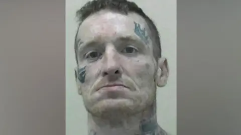 Northumbria Police Mugshot of Gary Haswell. He has short dark hair and a faint ginger stubble beard. He has various tattoos, including of a large crown above his left eye, a tribal mark around his right eye and stars and large flowers across his neck.