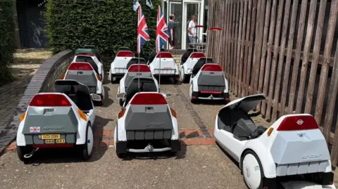 Simon Middleton About 10 C5 vehicles are parked in front of a building. Two of them have flags on them and people can be seen walking into the building