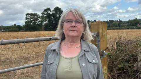 Charlie Stubbs / BBC There is a woman with grey shoulder-length hair. She is wearing a grey jacket and a light green T-shirt. She has a silver necklace and hoop ear-rings. She is stood in front of a metal gate which has barbed wire on the top. Behind her is a brown field. 