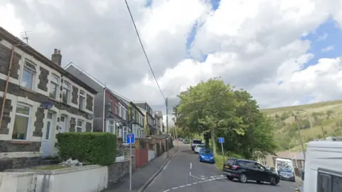Google street view images of Graig Road. it is a row of brick row houses on one side, raised from the pavement to a small front garden. the other side of the road is all tall leafy trees on a steep bank. A car is turning off the road down a steep incline. it's a cloudy day with some blue sky and the hills above the town can be seen in the background. 