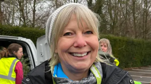 Tom Jackson/BBC Jenny Norton is in the middle. She has a blonde fringe and her hair is tied in a ponytail. She is wearing a grey hat, a black coat and a green lanyard around her neck. 
