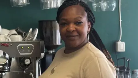 Bukola George, with long dark braided hair, tied back, standing in a cafe by a coffee machine. She is wearing a grey sweater and is looking at the camera and smiling.