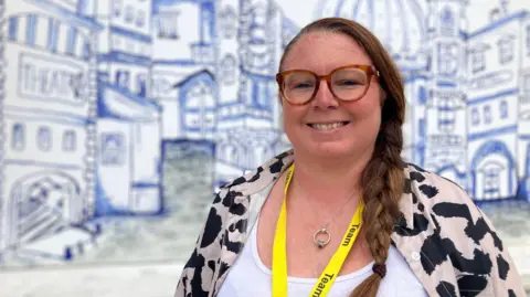 A woman wearing glasses, with her hair in a ponytail over her shoulder, smiles at the camera. She has a yellow lanyard around her neck and is front of blue and white artwork of buildings