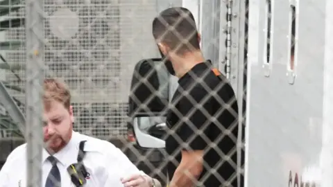 Eddie Mitchell a man wearing a black t-shirt facing away from the camera. He is being led out of a white prison van