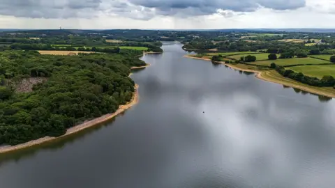 An aerial shot of a reservoir, with greenery on the banks and a grey cloudy sky in the distance