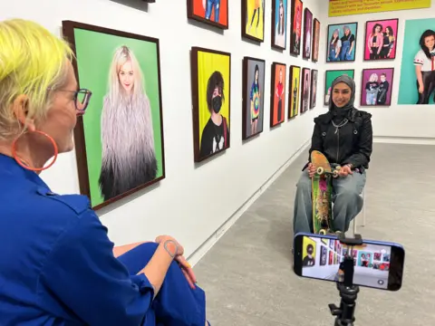 Casey Orr A young woman wearing a head covering sits in the middle of the exhibition holding her skateboard. She is facing the photographer 