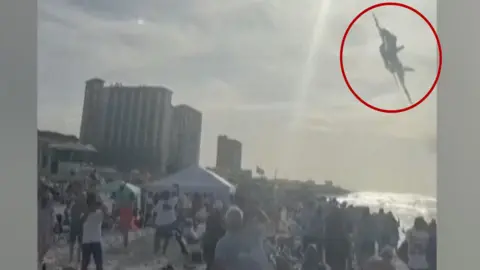 Jet flying above a crowd on a beach with a red circle around it.