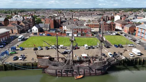 PA Media An aerial view of Blyth which sits on the water's edge. Rows of red brick buildings extend into the distance. A town square patches of grass in the middle and a number of cars parked up. There is a grand wooden boat moored up in the foreground.