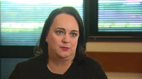 a woman looking at an off-screen interviewer. Her gaze is direct and she is wearing a black top and plum-coloured lipstick