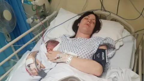 Supplied Kim lying in a hospital bed holding a new born baby and wearing a white gown. She has drips attached to her hands.