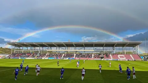 Joseph Perry Half-built football stand on match day with a rainbow in the sky above it
