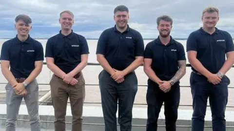 Five men, all in their twenties, are stood in a row smiling for the camera. They're stood on a balcony at the headquarters of their employer, the Spencer Group. All are wearing blue t-shirts, with the logo 'Spencer' on them and long trousers. All men have short hair and are smiling into the camera. The Humber Estuary can be seen behind them. 