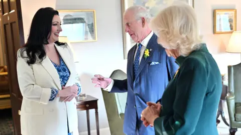 PA Media A dark haired woman in a white suit and blue shirt converses with a man in a blue suit and white shirt and a white haired woman in a green suit.  Behind them are gold framed paintings and two green leather chairs. The man has short grey hair and a yellow flower on his lapel. 