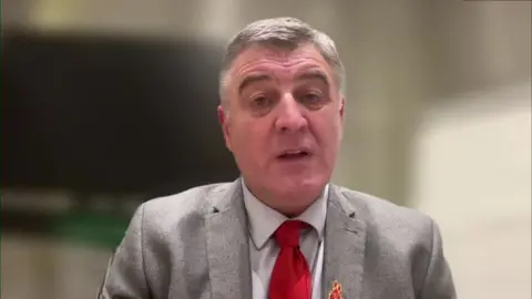 Ian has short grey hair, he wears a pale blue shirt under a grey blazer and a red tie 