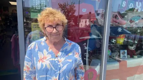 Annie Collins, a middle-aged woman with strawberry blonde hair, is wearing a blue flowery shirt and is standing in front of a charity shop window.