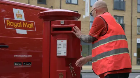 A postman unlocks a red post box in the street. His red Royal Mail is in the background 
