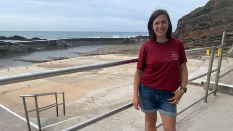 Jodie Harper stood to the right in front of the empty sea pool. She is wearing a red top and blue demin shorts. You can see the concrete at the bottom of the sea pool and Jodie is stood in front of silver railings. The sea is behind the walls of the pool.