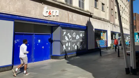 Alex Pope/BBC A B&M store with grey stone on the front, with a white B&M sign with a red &. There are several people walking past the store. The window frames are painted blue. There is a lamp post to the right. 