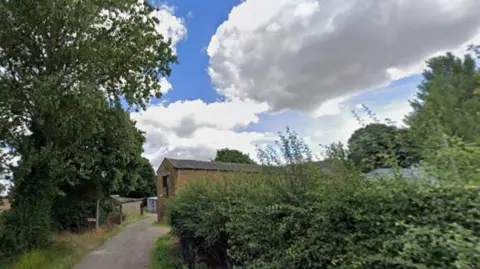 Google Streetview Alderthwaite Farm in Hoyland, Barnsley. A farm track with a hedge on the right, a corn field on the left, blue sky with white clouds, and an old barn at the end of the road.