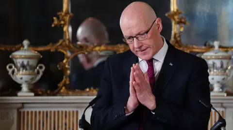 John Swinney in a dark suit, white shirt and burgundy tie holds his hands together as though praying. He is in front of a mirror.