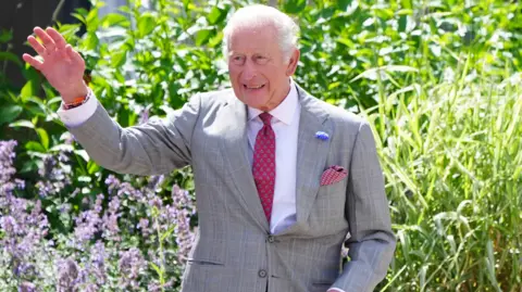 King Charles III is pictured wearing a grey suit, white shirt and red tie. He is waving.