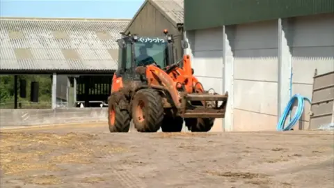 An orange tractor with a man in the cab drives towards a large farm building