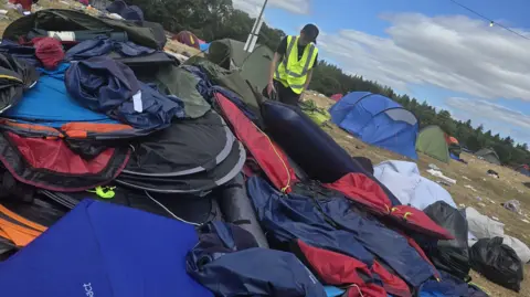 The Community Food Project A huge pile of discarded and colourful tents and air beds, in a large green field. in the background there are erected tents and rubbish strewn on the floor. A man with a black cap on and a yellow hi-vis jacket is standing over the pile