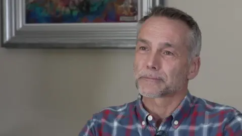 Lawrence Salvoni has short greying hair and a greying goatee beard. He wears a blue, red and white lumber shirt.