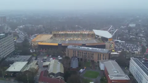 West Midlands Police An aerial view of Molineux stadium in Wolverhampton on an overcast day. The gold and black stands are visible along with surrounding car parks, roads and buildings. Police vehicles can be seen on the road below. 