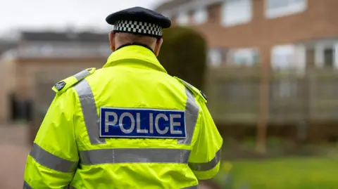 A police officer is walking away from the camera. He wears a yellow high vis coat, which has silver reflective panels down the sides and a rectangular logo along the centre which reads 'POLICE' on a blue background. He also wears a black cap with a black and white check panel around the head. The background is blurred but shows a housing estate.