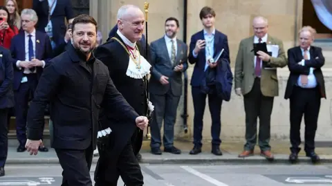 Getty Images Black Rod Ed Davis (centre) escorts Ukrainian President Volodymyr Zelensky during his visit to the Palace of Westminster on 17 March, 2026. He wears a black tailcoat with matching breeches, black stockings and slippers and a white ruffled collar. He carries his staff of office in his left hand.