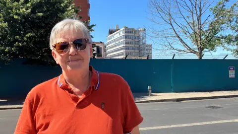 Owen Sennitt/BBC A woman wearing a sunglasses and a orange polo top looks towards the camera, with Sovereign House in view behind her