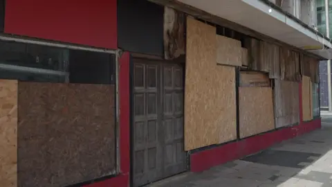 A red disused and rundown building. The photograph shows brown wooden doors in the middle of the building with its windows boarded up with yellow plywood.  Water damage can also be seen in the top of the building.