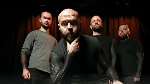 Donovan Jones Four members of Collapse The Sky look at the camera, three are standing and one is sitting down. They are all looking at the camera, unsmiling. They are wearing muted dark colours.