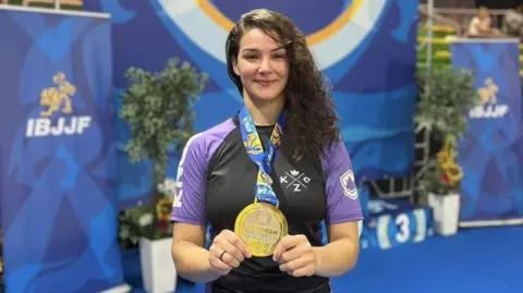 Bea Fernandes/Roots Gym Bea Fernandes holds up a medal towards the camera. She is dressed in a black top with purple sleeves.