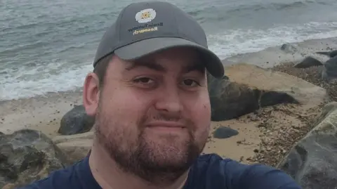 A man with a dark beard and moustache and green cap taking a selfie shot on a beach, There are large rocks scattered about with sea lapping at the shore.