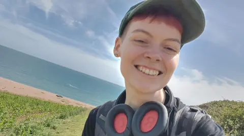 Name for Oxevision cameras to be banned in psychological well being sufferers' bedrooms Hat A woman stood on a hill with a beach and the sea behind her. She is smiling and has on a baseball cap and large headphones around her neck.
