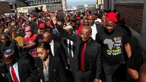 Reuters Julius Malema in a black suit and red tie walks out of court and waves at supporters who can be seen in the background.