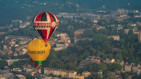 PA Media Two hot air balloons flying over Bristol. The city can be seen below, with the landscape featuring colourful properties and the Clifton Suspension Bridge as well as thick lines of trees.