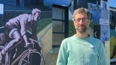 An adult stands in front of a large outdoor mural featuring a cyclist riding along railway tracks, a steam train, and dark birds flying overhead. Public toilet signs in blue and white are mounted on the mural wall behind the person.