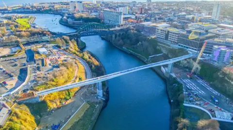 An aerial view of the footbridge. It is a long white structure crossing the River Wear and connecting Keel Square to the Stadium of Light. A crane is near it. Wearmouth Bridge and the North Sea are also visible in the distance.