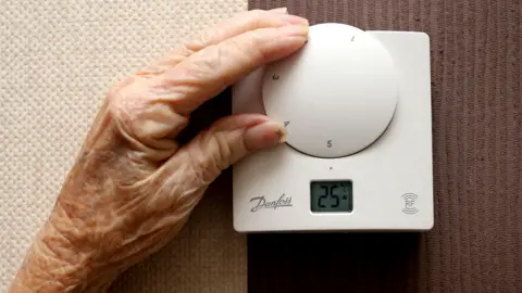 PA Media An older persons hand moves the dial on a white thermostat, the dial reads 25 degrees celcius