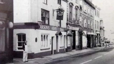 HCUK Black and white photograph of the Mason Arms building-with a blurry figure of a man standing next to it.
