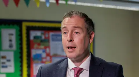 PA Media Paul Givan. He has short dark hair with white patches, wearing a dark blue blazer, white shirt and dark pink tie. Multi-coloured school notice boards are in the background.