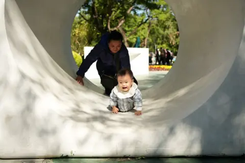 A baby crawls through a tunnel in the park, with an adult behind him